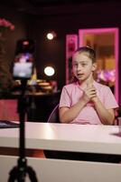 Gen Z entertainer utilizes smartphone on tripod to shoot introductory segment for vlog. Young internet star in pink neon studio welcomes devoted fan community, filming with phone photo