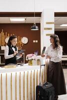 Elderly lady stands at hotel reception, where professional concierge assists with reservation. Senior woman completes check-in process with digital document on tablet. Suitcase is placed near counter. photo