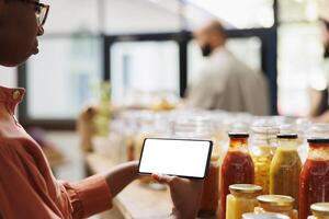 Black woman holding smartphone with blank white screen while looking at sustainable organic items. Consumer browses the counter for food products while gripping cellphone with chromakey display. photo