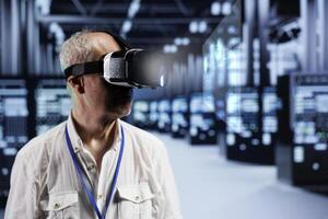 Old cloud computing business executive immersed in virtual reality at data center, doing equipment maintenance. Repairman using VR headset to optimize server rigs performance, checking code lines photo