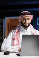 Image shows an Arabian man working at a desk, using a laptop and wireless technology. Muslim male embraces modern tech for his entrepreneurial endeavors by utilizing his personal computer. photo