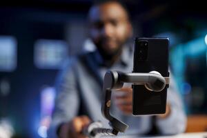 Enthusiastic internet show host in blurry background hitting the record button on cellphone. Focus on mobile phone attached to selfie stick used by content creator to record footage for fans photo
