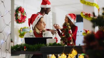 Woman filling paperwork in Christmas decorated office, perturbed by disturbing staff members discussing in background. Employee unable to concentrate on tasks while colleagues talk loudly photo