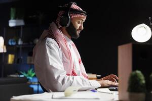 Side-view shot of a handsome Arab businessman works in modern office, using wireless technology and traditional attire. He engages in calls while browsing, researching, and communicating online. photo