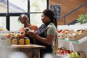 African american female vendor giving a complimentary treat to loyal customer at environmentally friendly market. Two black people talking about chemical free sustainable products at local store. photo