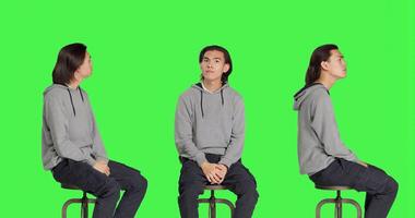 Young man sitting on chair and waiting for something in studio, looking at watch and searching for a thing against greenscreen backdrop. Male model with natural impatient expressions. photo