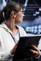 Old computer scientist updating data center rackmounts, writing intricate binary code scripts on tablet, using programming to fortify cybersecurity in technically advanced supercomputer hub photo