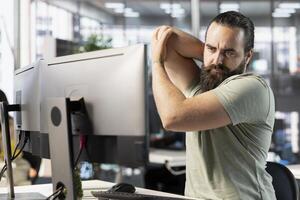 Startup employee suffering arm pain from bad posture on office chair while doing research for project. Man feeling muscle strain, stretching while looking at computer in workspace, solving tasks photo