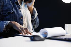 Close-up of black individual using smartphone to talk while studying documents at a desk. African American freelancer with her hand on a clipboard as she checks and examines papers, working from home. photo