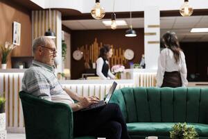 Female guest checks-in at hotel reception while eldery male tourist browses and does room booking using laptop. Retired senior man using his personal computer for online check-in. photo
