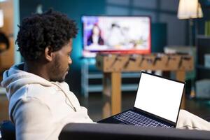 Self employed man inputting data on mockup laptop display while working from home, looking at screen. Person lying on couch, remotely performing tasks on isolated screen notebook in apartment photo