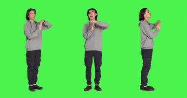 Model does timeout symbol with hands, asking for a break or pause against greenscreen in studio. Asian man showing disapproval and negative emotions on camera, stop gesture. photo