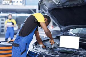 Mockup laptop on car with hood open while repairman in blurry background replaces motor. Isolated screen device next to licensed garage specialist fixing customer automobile photo