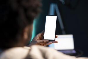 Cozy african american man chatting with online friends using mockup phone in warm stylish home, enjoying himself. Person scrolling on isolated screen cellphone entertained by social media activity photo