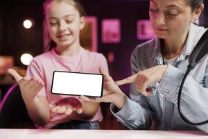 Happy child and mother filming assessing mockup smartphone performance, talking about components used. Daughter and parent shoot isolated screen mobile phone review for online channel photo