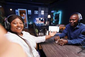 African lady filming podcast with guest telling listeners an engaging narrative using selfie camera on cell phone. Female interviewer introducing male influencer to online audience using mobile device photo