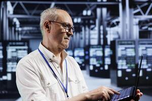 Elderly technician using laptop to examine blade servers for power fluctuations. Efficient professional monitoring data center hardware, identifying potential vulnerabilities photo