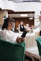 Detailed view of elderly lady client relaxing in cozy lounge area with tablet and wireless headset. Retired old woman listening to music and browsing internet on digital device. Close-up, side-view. photo