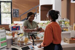 Black woman in apron assists customer at food market counter. Emphasis on local, organic produce promotes healthy, eco-friendly lifestyle. Small business owner and shopper support community together. photo