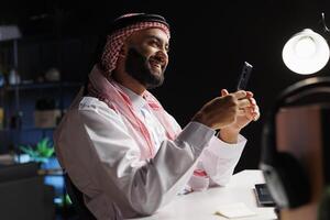 Side-view of smiling Muslim man in professional attire works at a modern desk. Using a smartphone, he browses the internet, representing the digital age and wireless technology. photo