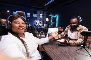 Smiling radio host holding a mobile device and taking a selfie with social media star in home studio. African american content creators doing a podcast episode and vlogging with smartphone. photo