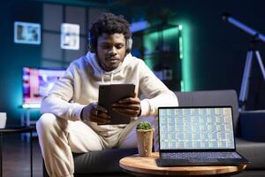 Man listens to music and reads from tablet, waiting for stock market financial figures on laptop screen to get updated. Businessman at home next to notebook showing trading investment charts photo