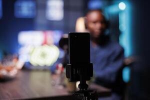 Focus on smartphone on tripod recording vlogger show host in blurry background interviewing guest in neon lights studio. Detailed view of mobile device being used to film content for male influencer. photo