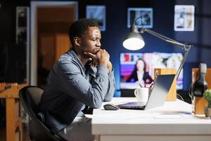 Stressed employee trying to solve executive responsibilities from home, doing remote work at night. African american worker reading management strategy plan to help with development. photo