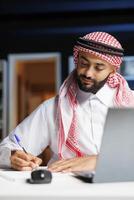 A focused Middle Eastern man working at a table with a notebook is shown in detail, dressed traditionally. A close-up of a Muslim man writing out research at his desk at home. photo