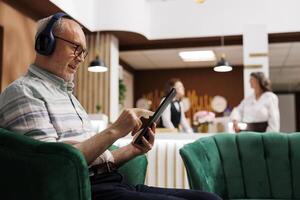 Retired senior man sits on couch in lounge area surfing the web on device. Elderly guy relaxes on comfortable sofa using tablet to make online reservation and listens to music with headphones. photo