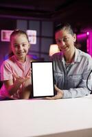 Happy child and mom filming assessing mockup tablet performance, talking about components used. Daughter and parent shoot isolated screen digital device review for online channel photo