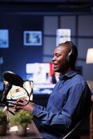 Cheerful african guy in side view profile is seen speaking into modern microphone while wearing a wireless headset. In home studio, male content creator is using audio equipment to record a talk show. photo