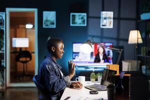 African american blogger waving at her personal computer while seated at a desk having a call. Black woman uses digital laptop to chat online and research while immersed in digital technology. photo
