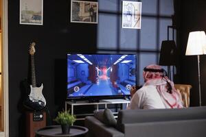 Image showing a Middle Eastern man in traditional clothing concentrated on an intense online game. Arab guy seated on the couch using wireless controller, having some fun digital entertainment. photo