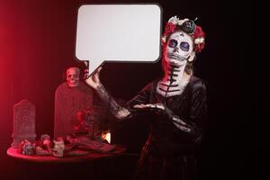 Woman acting as goddess of death holding speech bubble, wearing skull makeup for dios de los muertes event. Woman using isolated copyspace template on cardboard, santa muerte advertisement photo