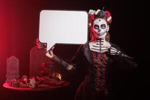 Portrait of woman dressed in Day of the Dead costume celebrating Dia de los Muertos, holding speech bubble. Girl wearing skeleton makeup using isolated copyspace template, promoting Mexican culture photo