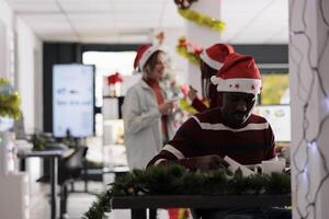Exhausted african american employee stressed about incoming task deadline during Christmas season. Worker multitasking in festive themed office while colleagues in merry holiday mood enjoy themselves photo