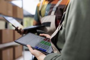 Stockroom colleagues analyzing inventory report on tablet computer, working at clients orders before shipping packages in warehouse. Diverse supervisors looking at merchandise checklist. Close up photo
