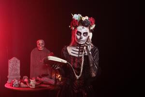 Woman using landline phone to answer call in graveyard, celebrating Day of the Dead. Mysterious lady talking on office cord telephone surrounded by frightening funeral items during Halloween event photo