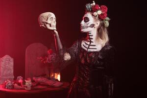 Woman posing with skull in hand, acting Hamlet scene while wearing skeleton makeup. Macabre lady dressed as La Catrina character on day of the dead event in studio background with red light photo