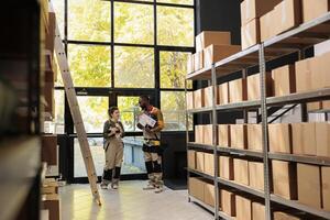 Diverse stockroom team looking at cardboard boxes checking clients orders in storehouse preparing packages for delivery. African american employee wearing overall while working at warehouse inventory photo