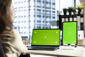 Business director working in her own office with green screen, looking at device display and developing a new project idea. Female entrepreneur empowered by corporate culture. photo