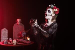 Woman raging after losing videogame during Day of the Dead. Mysterious lady holding controller, upset after being defeated, surrounded by frightening funeral items during Halloween horror event photo