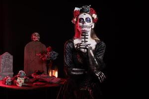 Portrait of woman during Day of the Dead Mexican event honoring dead loved ones, graveyard background. Scary lady with cadaver body paint during ancient Aztec custom of celebrating ancestors photo