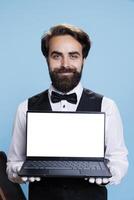 Young bellboy holds white display on laptop in studio while he wears formal suit and tie next to luggage. Classy doorkeeper working at luxurious hotel and shows blank screen template. photo