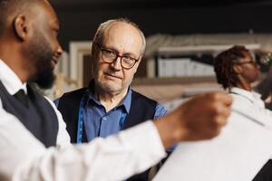 Close up of master tailor teaching skillful craftmanship to aspiring fashion design apprentices in atelier workspace. Diverse team of couturiers working together on bespoken sartorial garments photo