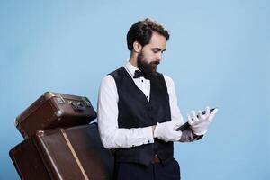 Hotel employee looking over bookings to make online reservations on tablet, working in hospitality and tourism industry. Professional bellhop reads list of guests, posing with luggage in studio. photo
