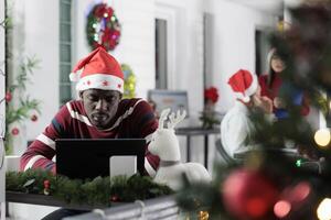 Sleepy businessman in festive adorn office working on demanding project after sleepless night. Employee struggling to remain awake while solving tasks at work during Christmas holiday season photo