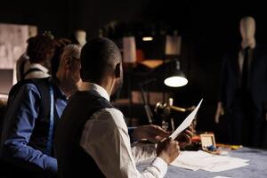 African american client discussing over clothing sketches with skilled craftsman, asking for customization alterations to be done. Suitmaker helping gentleman with bespoke attire in atelier studio photo