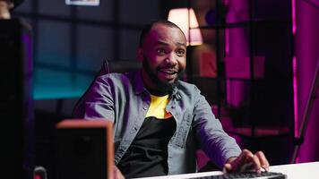 Joyous gamer competing in intense internet multiplayer videogame from vibrant living room with neon lights. Smiling african american man using PC, having immersive experience while gaming, camera A photo